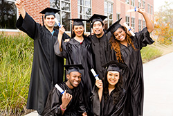 Six young adults in black cap and gowns smiling and holding their diplomas