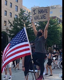 masked black woman at protest holding up I Have Value sign next to American flag