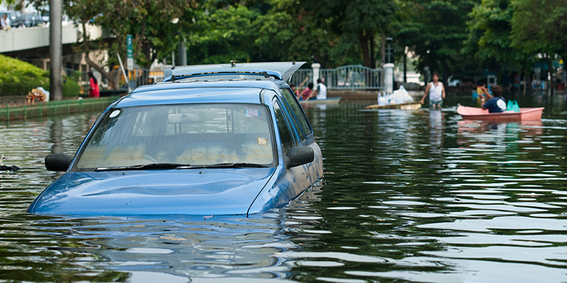 Blue car stuck in flooded road with water up to the headlights and people in the background