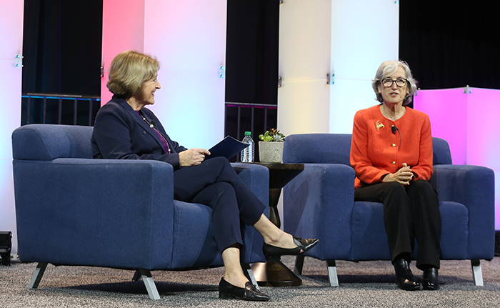 Two white women sit in blue chairs and are in conversation for attendees at the APHA Opening Session