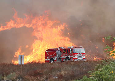 A firetruck sits in the foreground of a wildfire sweep across hills